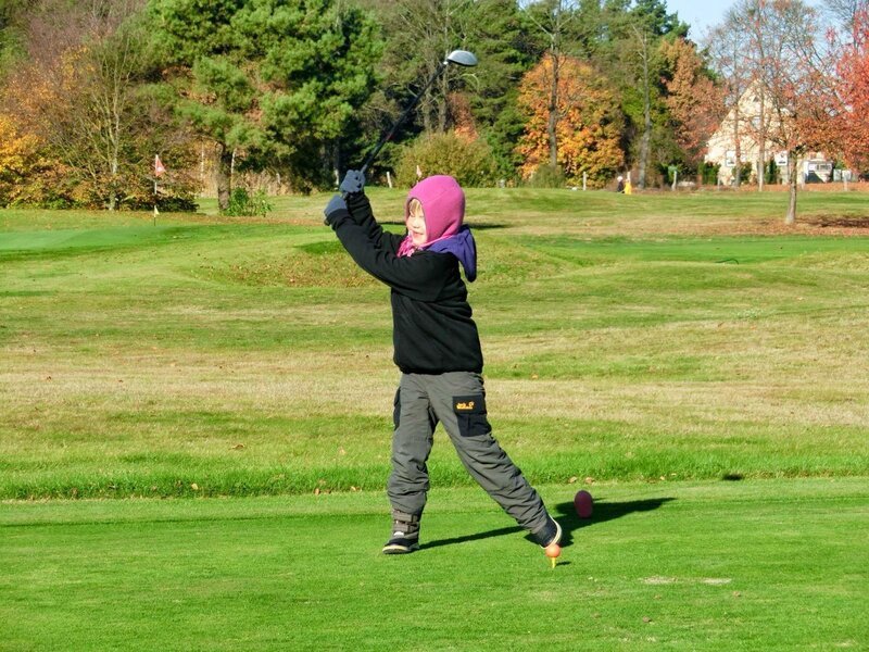Young child golfer swinging a club on a golf course in autumn, bundled up in warm clothes.