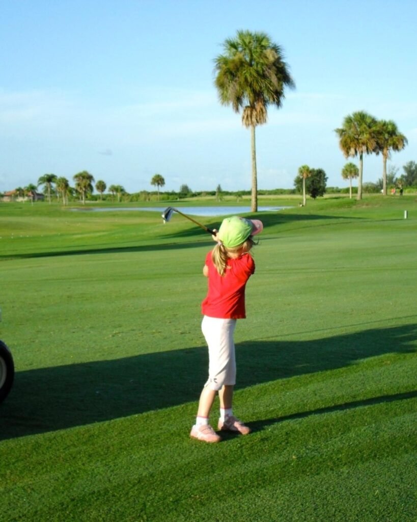 Young Keira Wolf swinging a golf club on a sunny course with palm trees in the background.
