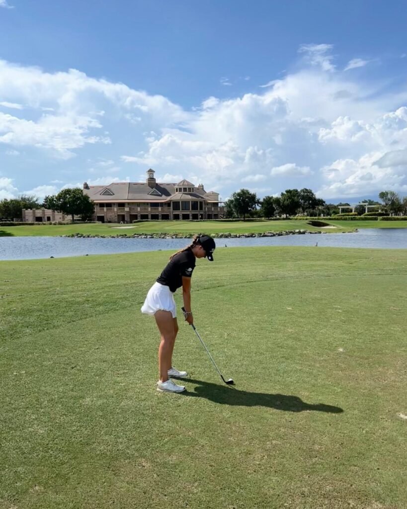 Keira Wolf preparing to hit an iron shot during an international junior golf tournament with a clubhouse and lake in the background.