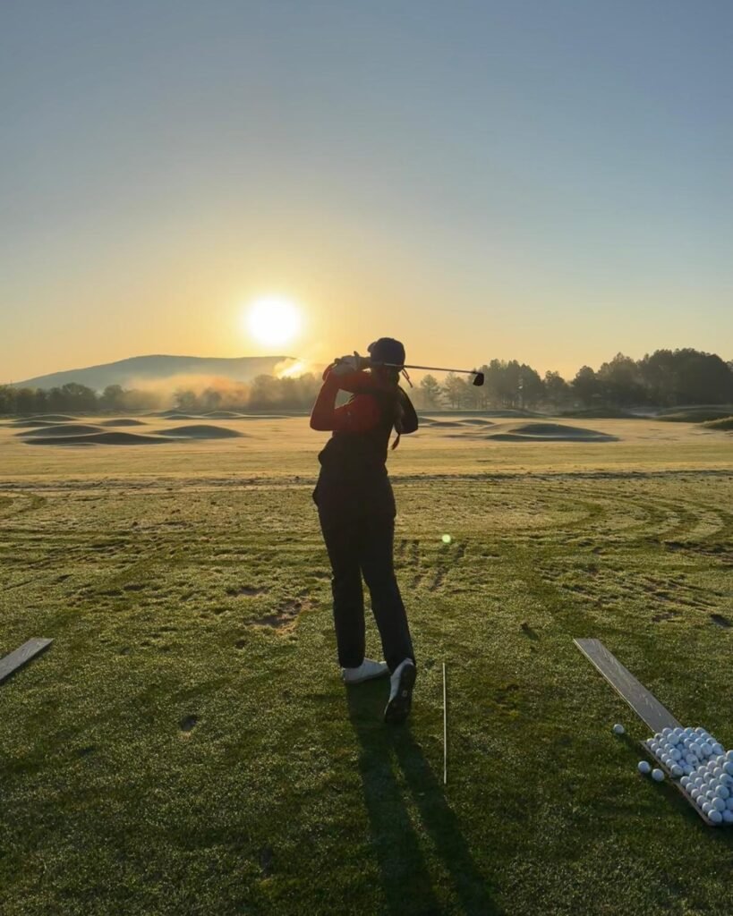 Keira Wolf hitting a golf shot on the driving range at sunrise with practice balls on the ground.