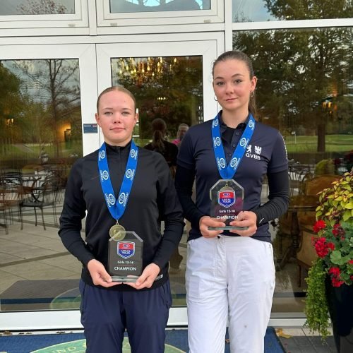 Two junior female golfers standing with trophies and medals in front of a golf clubhouse after winning youth championship categories Girls 13–14 and Girls 15–18