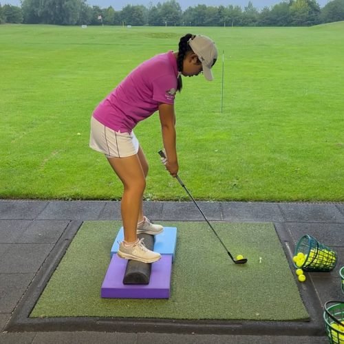 Female golfer on driving range standing on balance pads and half foam roller while practicing swing with training balls