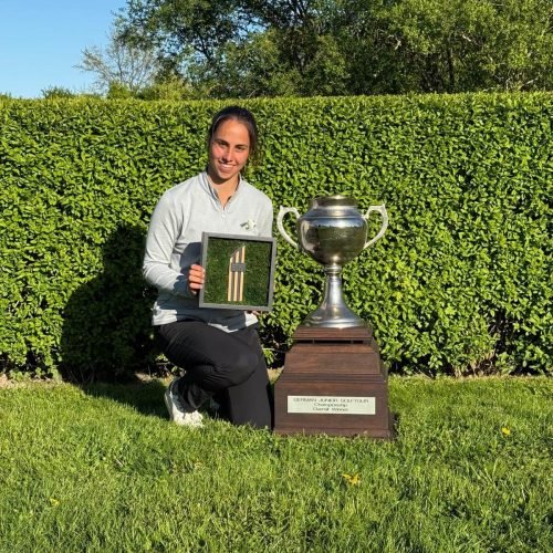 Female golfer kneeling on grass holding an award beside the German Junior Golf Tour championship trophy