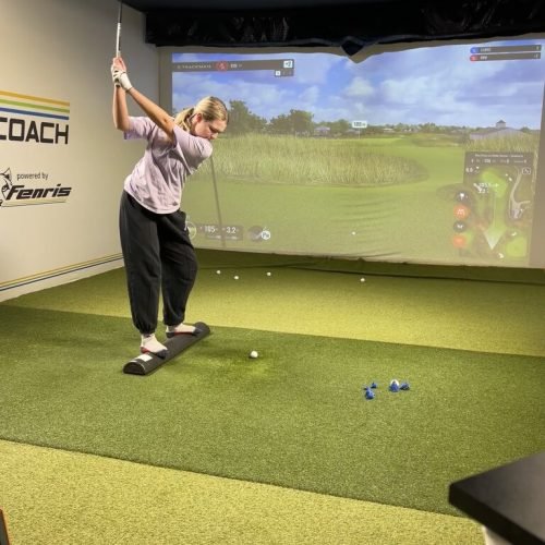 Female golfer swinging indoors on a TrackMan simulator with digital golf course projection and shot data on screen