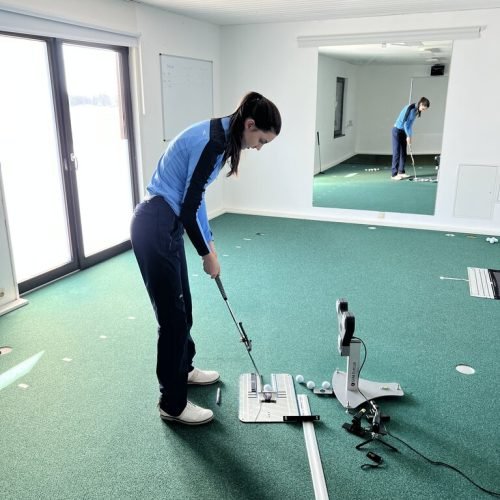 Female golfer practicing putting indoors with analysis equipment, training aids, and mirror reflection